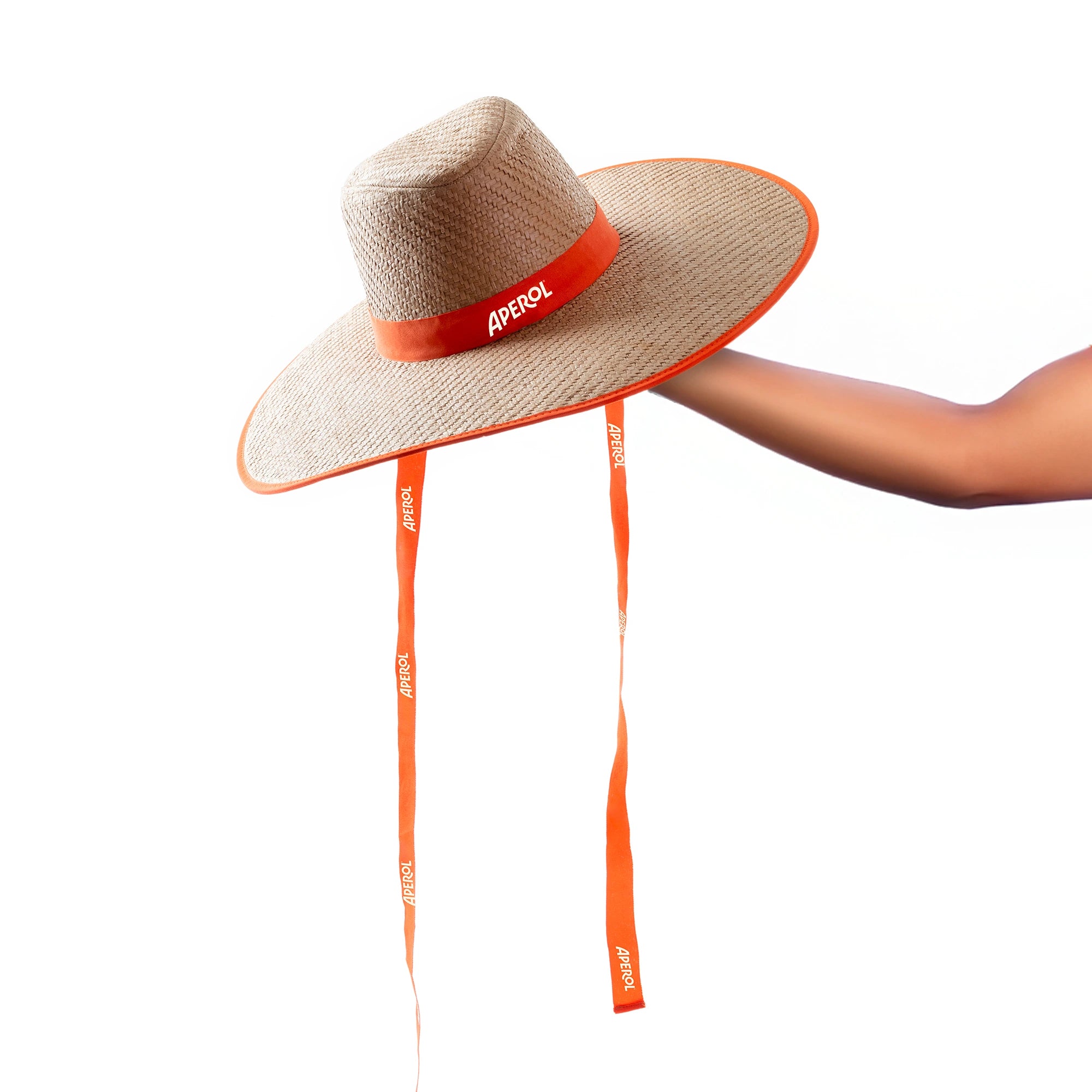 Wide-brimmed straw hat with an orange Aperol-branded ribbon around the crown and two long orange chin straps, set against a white background.